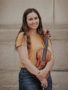 Hayley Camp stands, facing the camera and holding her violin.